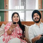 Tunic Styling. - Cheerful wife with bindi on forehead wearing plaid tunic with white trousers using TV remote control for channel switching while lying on bed with laughing Sikh husband in turban with stylish beard and twisted mustache